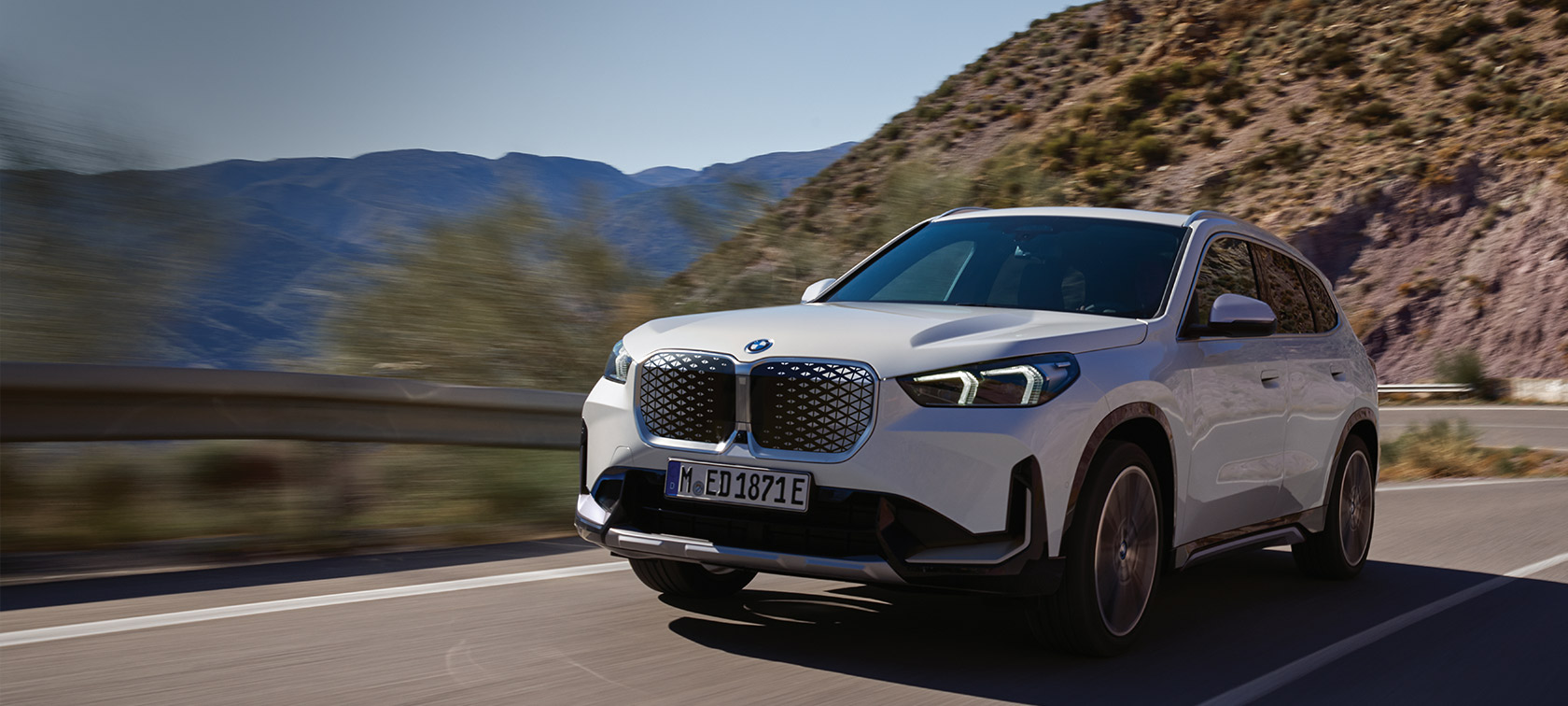 BMW iX1 U11 BEV 2022 Mineral White metalizado, vista de tres cuartos del frontal, en marcha por una sinuosa carretera de montaña